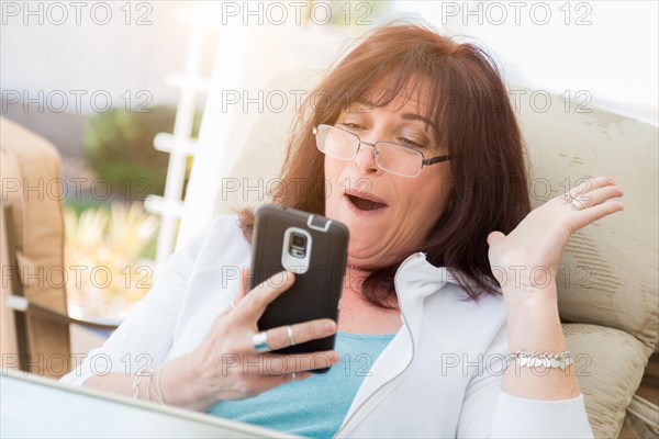 Shocked middle aged woman gasps while using her smart phone on the patio