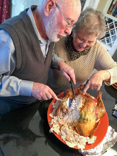 Senior adult couple cutting the holiday Turkey together in the kitchen