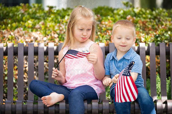 Young sister and brother comparing each others american flag size on the bench at the park