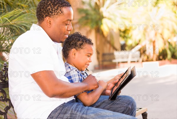 African american father and mixed-race son using computer tablet on bench in park