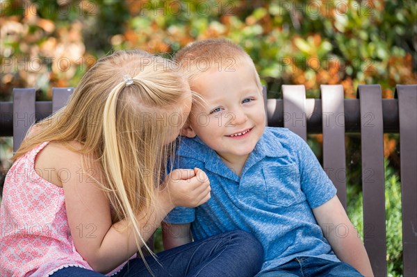Young sister and brother whispering secrets on the bench at the park