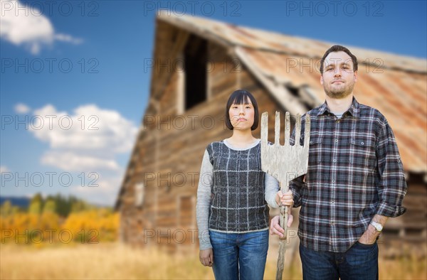 Biracial chinese and caucasian young couple holding wooden pitch fork in front of rustic barn in the country