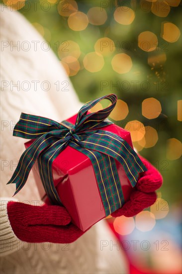 Woman wearing red mittens holding christmas gift against decorated tree and lights