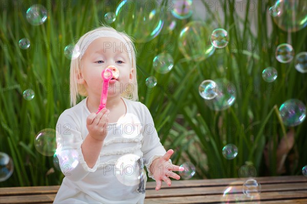 Adorable little girl sitting on bench having fun with blowing bubbles outside