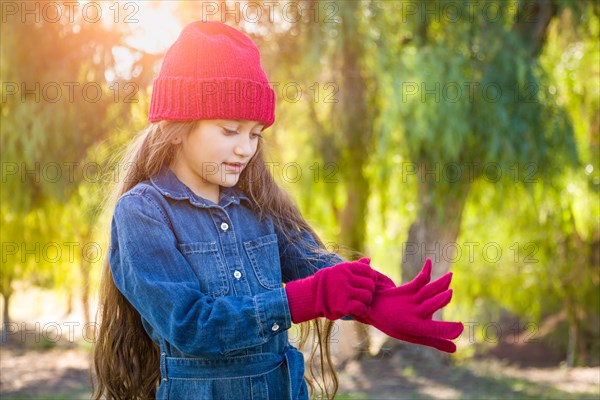 Cute mixed-race young girl wearing red knit cap putting on mittens outdoors