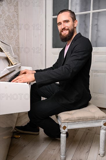 An einem Klavier sitzt ein junger Mann mit Vollbart und kurzen Haaren. Er blickt zur Kamera waehrend er beide Haende auf den Tasten liegen hat. Der schwarze Anzug weist den Spieler als Profi aus