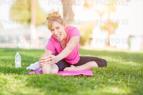 Young fit flexible adult woman outdoors on the grass with yoga mat stretching her legs