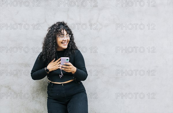 Young latin woman happy with her cell phone leaning against a wall. A young latin woman laughing with her cell phone leaning against a wall