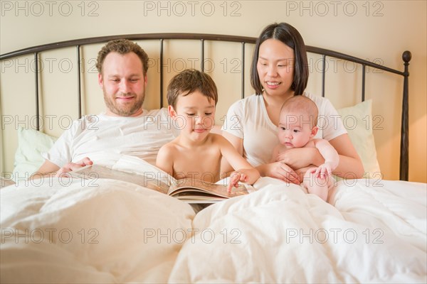 Young mixed-race chinese and caucasian baby boys reading a book in bed with their father and mother