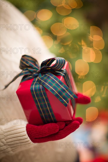 Woman wearing red mittens holding christmas gift against decorated tree and lights