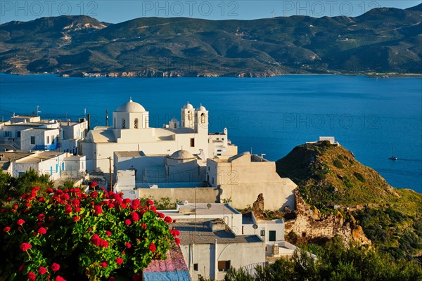 Picturesque scenic view of Greek town Plaka on Milos island over red geranium flowers and Orthodox greek church