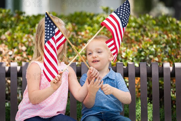 Young sister and brother waving american flags on the bench at the park