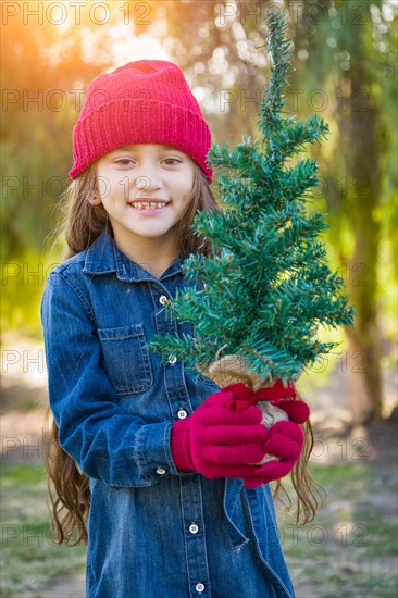 Cute mixed-race young girl wearing red knit cap and mittens holding small christmas tree outdoors