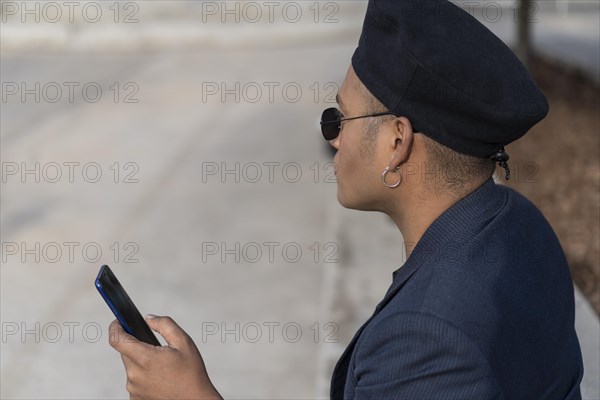 Latino gay male with makeup on wearing trendy hat holding a cellphone in a park