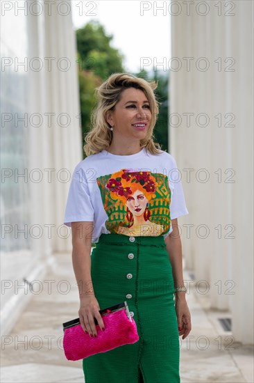 Portrait in einem Park einer huebschen Dame in ihren vierziger Jahren mit langen blonden Haaren. Sie geht auf die Kamera zu und blickt mit ueberraschte Blick zu Seite. Ein gruener langer Rock mit bedrucktem Shirt ergibt ein elegantes Outfit. In der rechten Hand traegt sie eine rosa Clutch