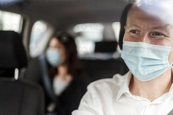 A happy taxi driver wearing a mask and the passenger on the back seat of the car in the background