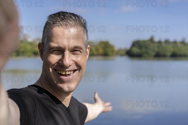 Active handsome mature Caucasian man taking selfie showing around at a lake. Outdoor adventure concept