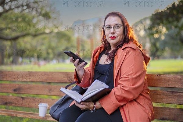 Woman sitting on a bench in a park checking her agenda and cell phone while having a coffee