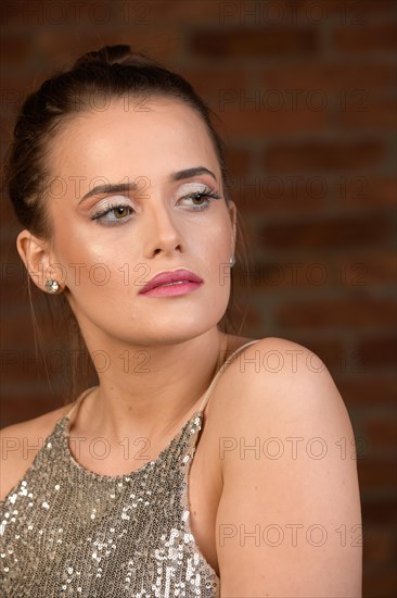 Huebsche junge Frau mit langen Haaren und dezentem Make-Up vor braunem Hintergrund. Das glitzernde Shirt passt zum dezenten Make-Up. Der Blick ist ueber die freie Schulter nach hinten an der Kamera vorbei gerichtet