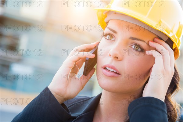 Young worried looking professional female contractor wearing hard hat at construction site using cell phone