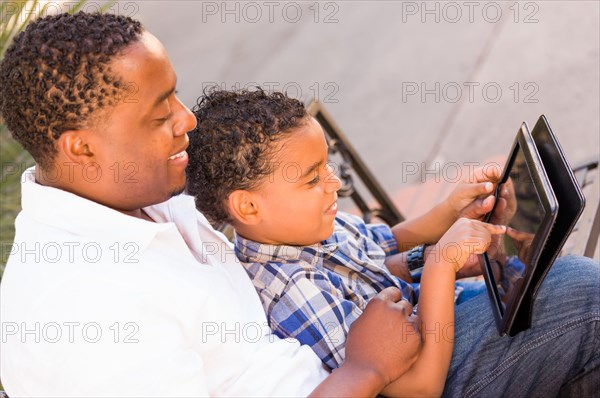 African american father and mixed-race son using computer tablet on bench in park