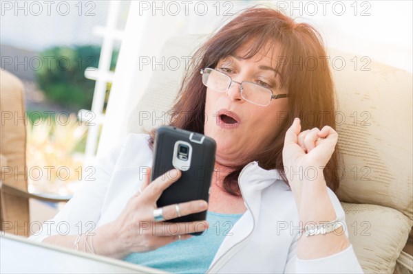 Shocked middle aged woman gasps while using her smart phone on the patio