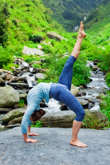 Woman doing yoga asana eka pada urdva dhanurasana Upward Bow Pose outdoors at waterfall in Himalayas