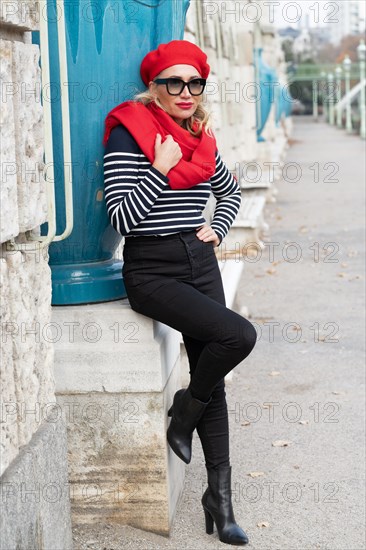 In einem Park lehnt an einer Mauer eine gut aussehende Dame mit roter Muetze und passendem Schal. Die schwarze Hose mit den Stiefeln passt zum Pullover im Matrosenlook. Die grosse schwarze Sonnenbrille ist ein guter Kontrast zu den blonden langen Haaren. Vienna