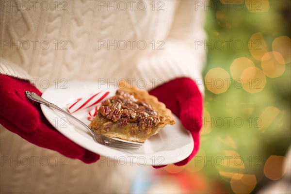 Woman wearing red mittens holding plate of pecan pie with peppermint candy against decorated tree and lights