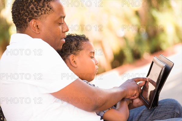 African american father and mixed-race son using computer tablet on bench in park