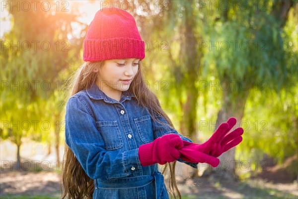 Cute mixed-race young girl wearing red knit cap putting on mittens outdoors