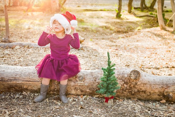 Cute mixed-race young baby girl having fun with santa hat and christmas tree outdoors on log