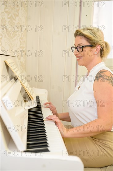 An einem Klavier sitzt eine attraktive junge Frau mit langen blonden Haaren. Die aermellose weisse Bluse und der beige Rock passen zu dem hellen Ambiente dieses Raumes