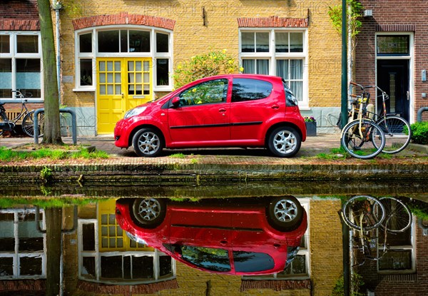 Red car on canal embankment in street of Delft with reflection and bicycle