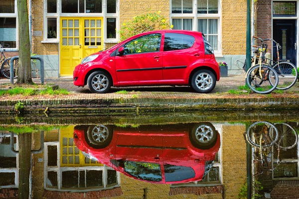 Red car on canal embankment in street of Delft with reflection and bicycle