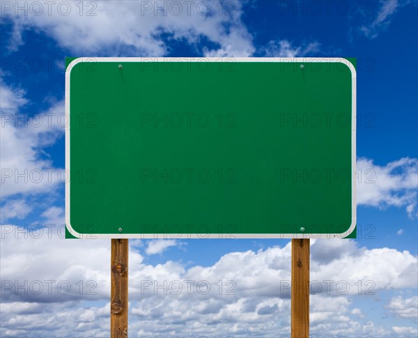 Blank green road sign with wooden posts over blue sky and clouds