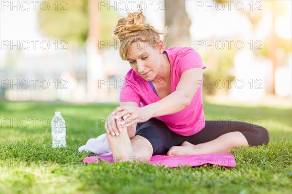 Young fit flexible adult woman outdoors on the grass with yoga mat stretching her legs