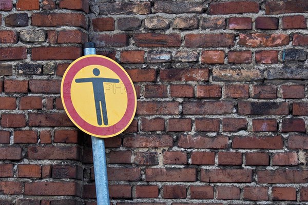 Round traffic sign with man gesturing with outstretched arms in front of brick wall