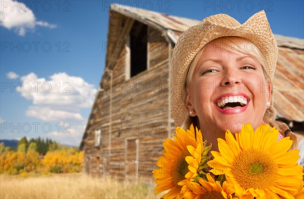 Beautiful caucasian young woman wearing cowboy hat holding sunflower in front of rustic barn in the country