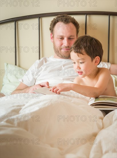 Chinese and caucasian baby boy reading a book in bed with his daddy