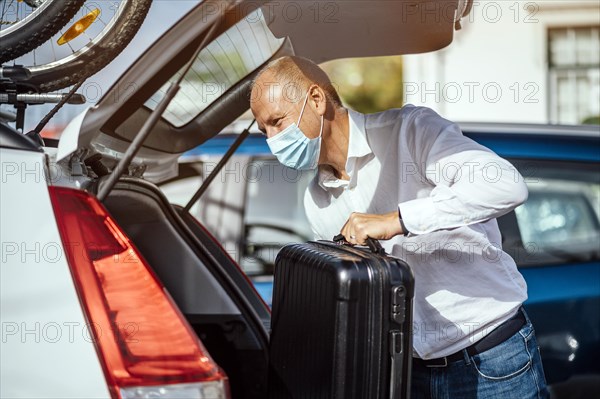 A taxi or Uber driver in mask puts the luggage into the trunk of his car