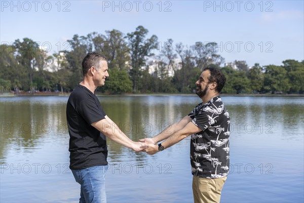 Homosexual mature white male couple holding hands smiling at a lake