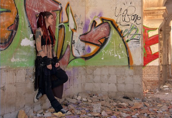 Young woman with tatoos and dreadlocks stands against wall with graffiti of an abandoned house