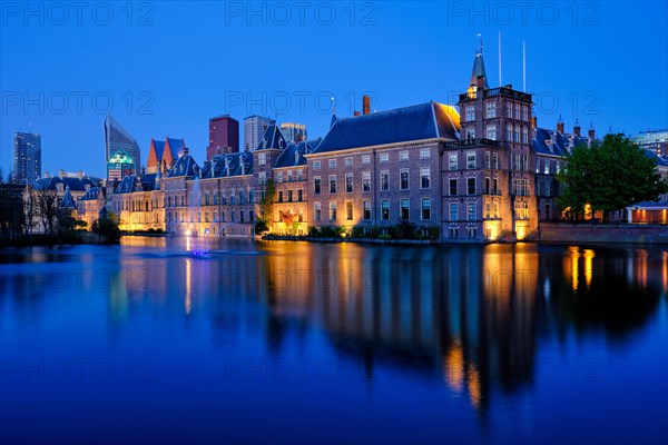 View of the Binnenhof House of Parliament and the Hofvijver lake with downtown skyscrapers in background illuminated in the evening. The Hague