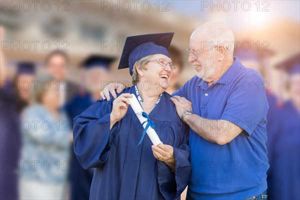 Senior woman in hat and gown being congratulated by husband at outdoor graduation ceremony