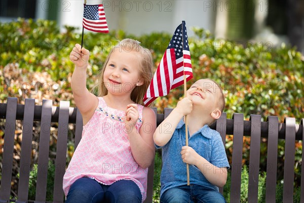 Young sister and brother comparing each others american flag size on the bench at the park