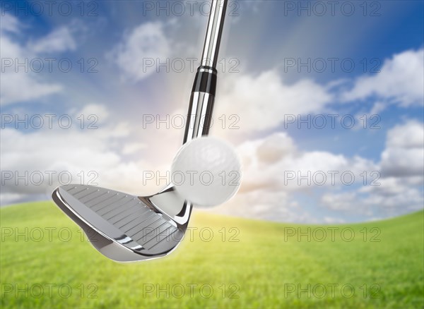 Chrome golf club wedge iron hitting golf ball against grass and blue sky background