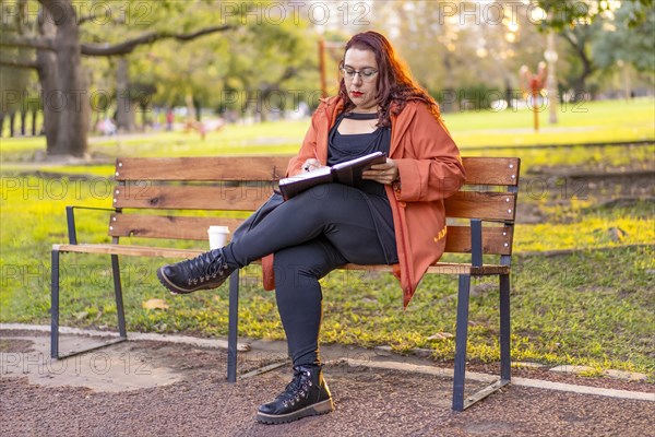 Woman sitting on a bench in a park checking her agenda and cell phone while having a coffee