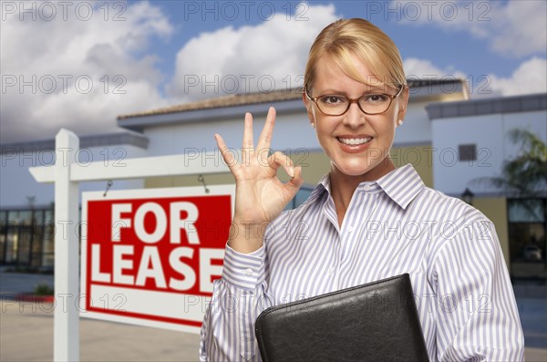 Smiling businesswoman with okay sign in front of vacant office building and for lease real estate sign