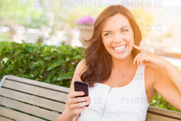 Attractive smiling young adult female texting on cell phone outdoors on a bench